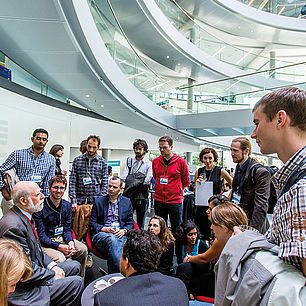 A photograph of a group of young researchers, standing in a modern building, grouped around a seated old bearded internet evangelist